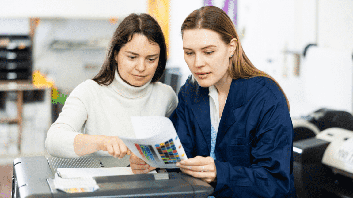 Two women review a colour chart amidst printing equipment.